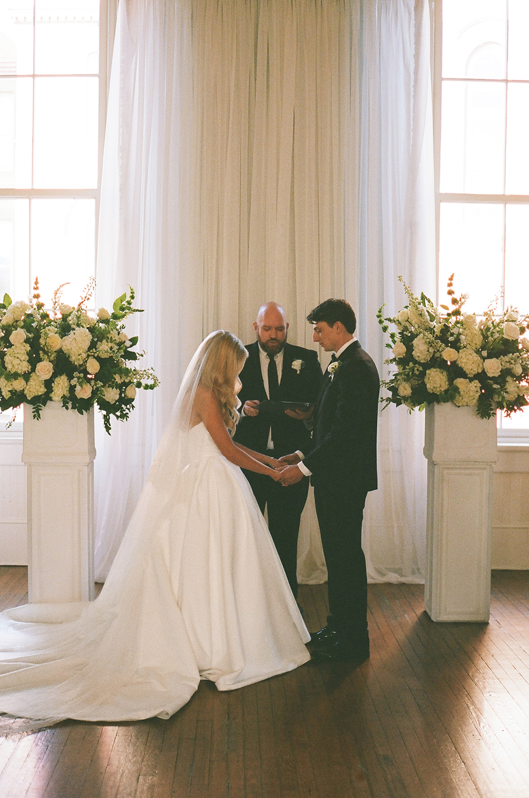 A bride and groom kiss indoors in front of tall windows, flanked by two large white floral arrangements on pedestals at the Stanly Ballroom a wedding venue in North Carolina