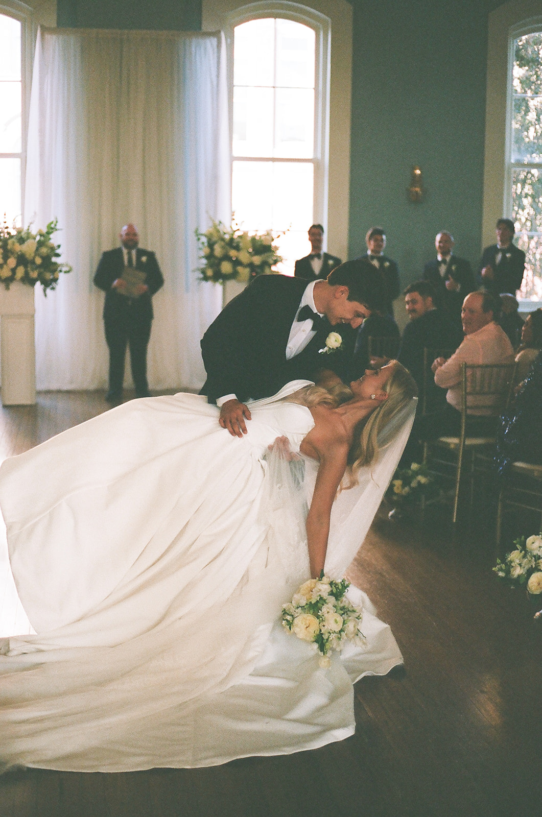 A bride and groom kiss indoors in front of tall windows, flanked by two large white floral arrangements on pedestals at the Stanly Ballroom a wedding venue in North Carolina