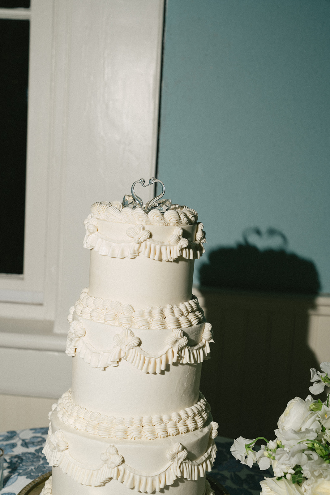 Three-tiered white wedding cake with ornate icing decorations and a small heart-shaped topper, placed on a table against a blue wall.