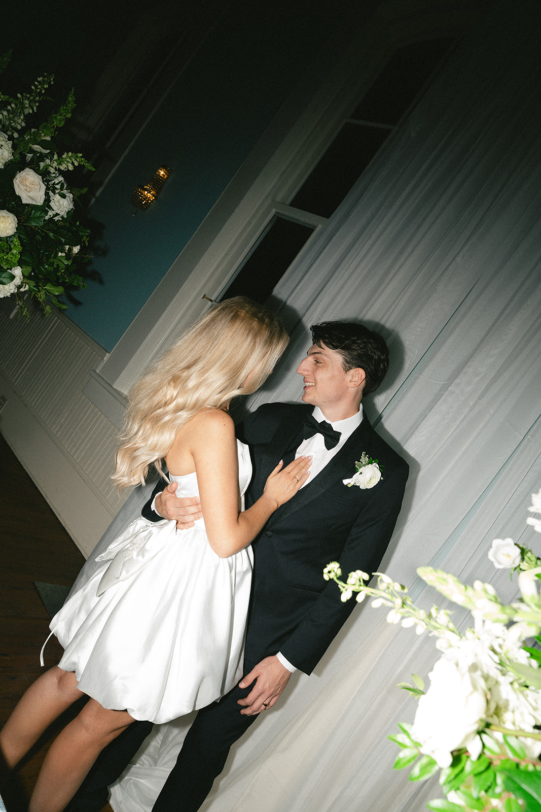 A couple dressed in formal attire, the woman in a white dress and the man in a black tuxedo, stand close together indoors, surrounded by white floral arrangements.