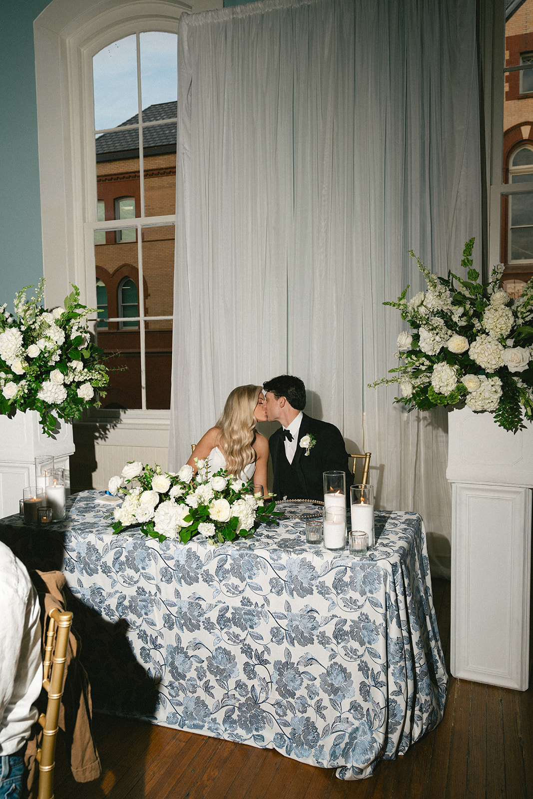 A couple dressed in formal attire, the woman in a white dress and the man in a black tuxedo, stand close together indoors, surrounded by white floral arrangements.
