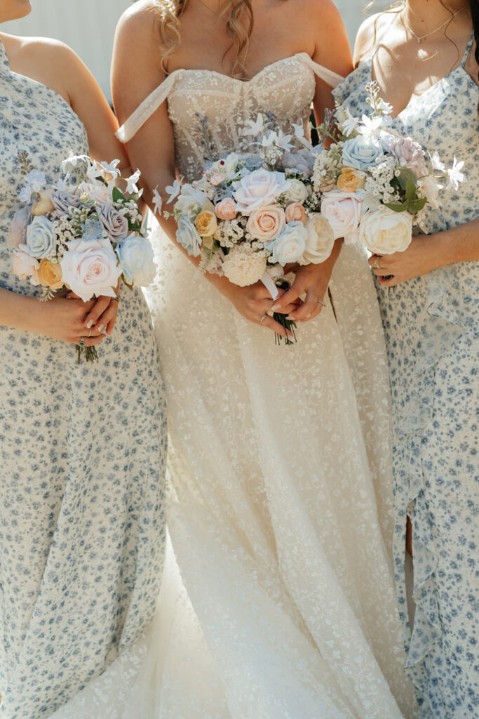 Three women in light floral dresses hold pastel flower bouquets; the woman in the center wears a white, off-the-shoulder wedding gown.