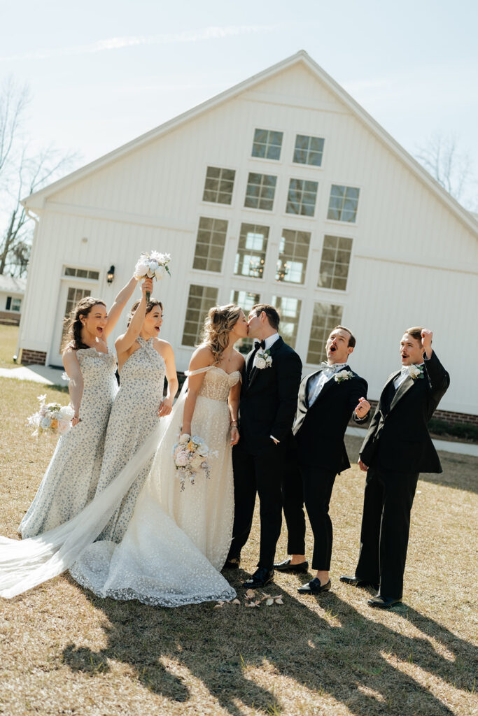 Two brides and three groomsmen stand outside a white building; the brides celebrate with bouquets while one kisses a groom, all dressed in formal wedding attire.