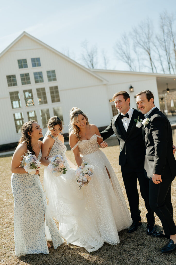 Two brides and three groomsmen stand outside a white building; the brides celebrate with bouquets while one kisses a groom, all dressed in formal wedding attire.