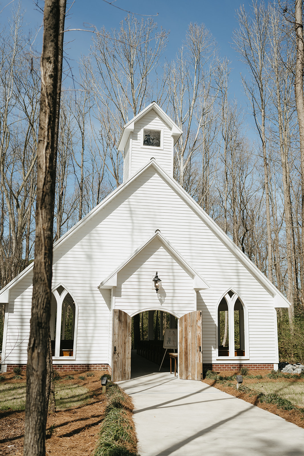 A small white chapel with wooden doors sits among tall leafless trees on a clear day, with a concrete path leading to the entrance at Birdsong Chapel 
