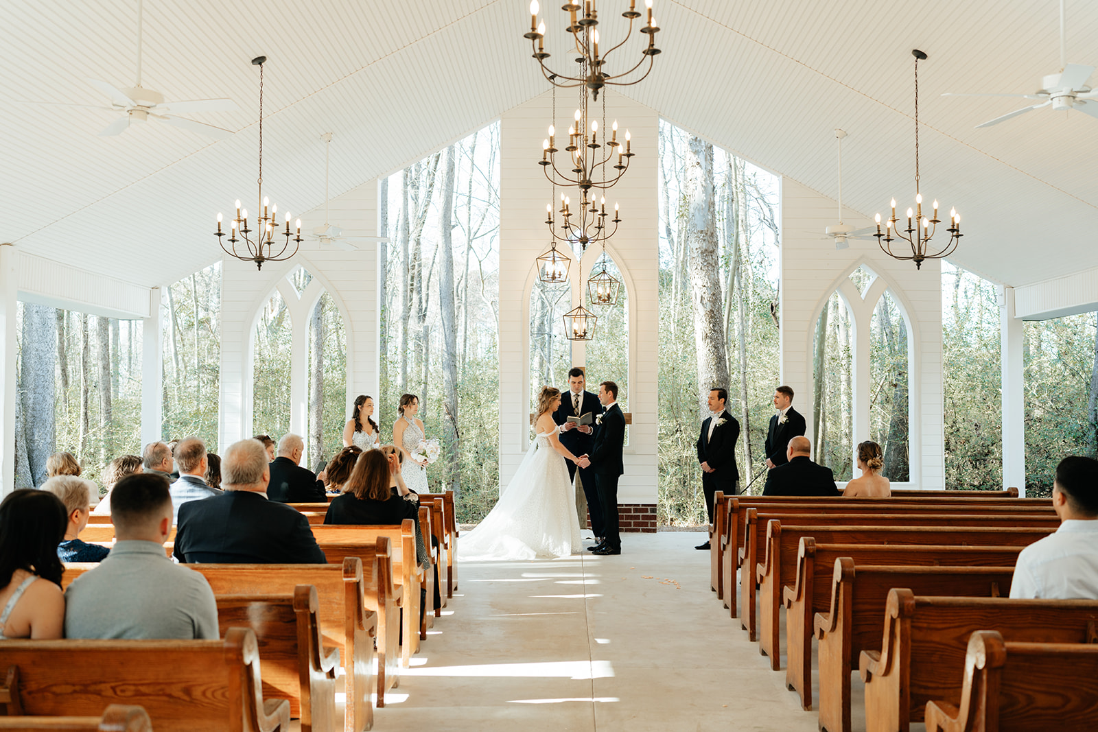 A wedding ceremony takes place inside a bright chapel with large windows, wooden pews, and guests seated facing the couple at the altar.