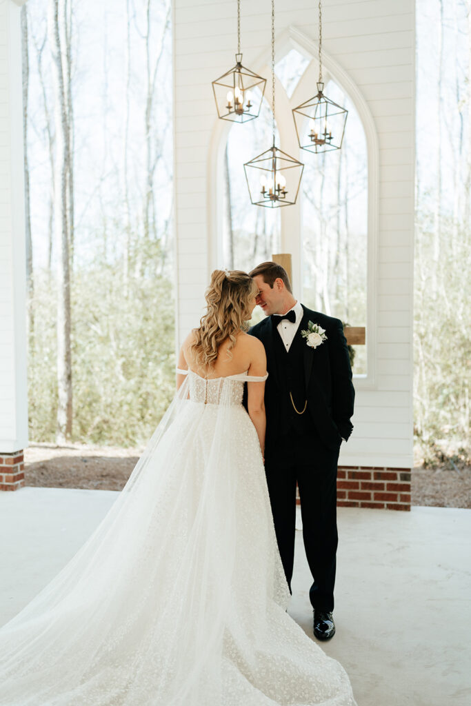A bride in a white gown and a groom in a black tuxedo stand close together in a bright, airy chapel with large windows and hanging lanterns.
