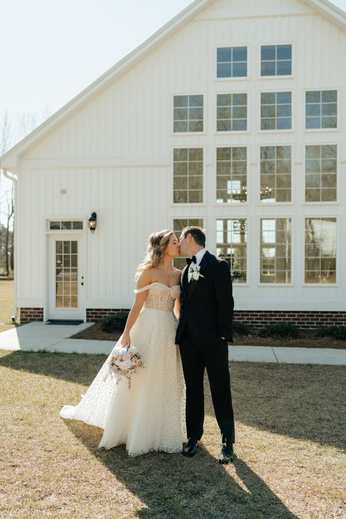 A bride and groom stand outside a white building, sharing a kiss. The bride holds a bouquet and wears a white dress; the groom is in a black suit.