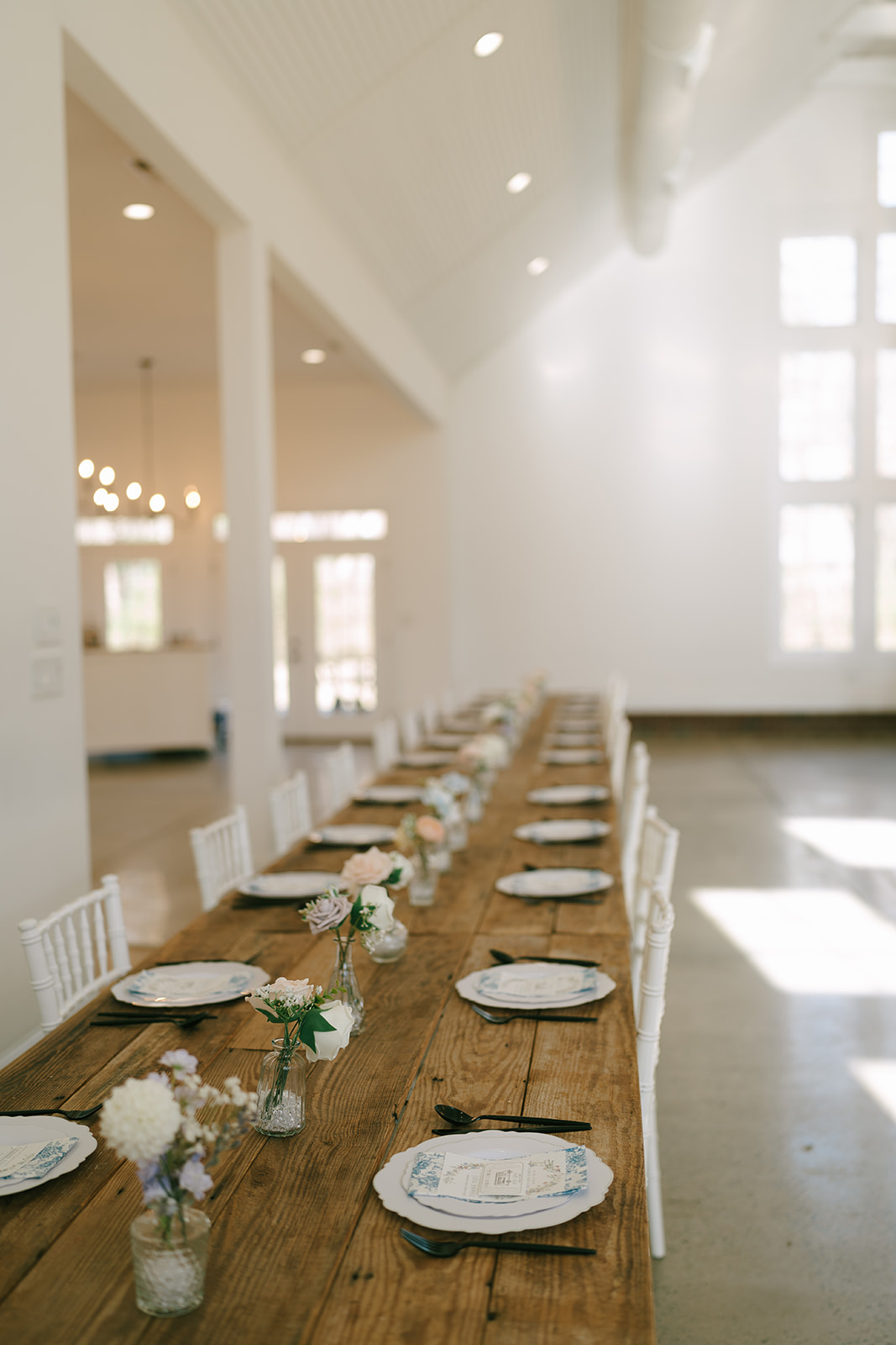 A long wooden table with white chairs is set in a bright, airy room with large windows, high ceilings, chandeliers, and minimal decor.