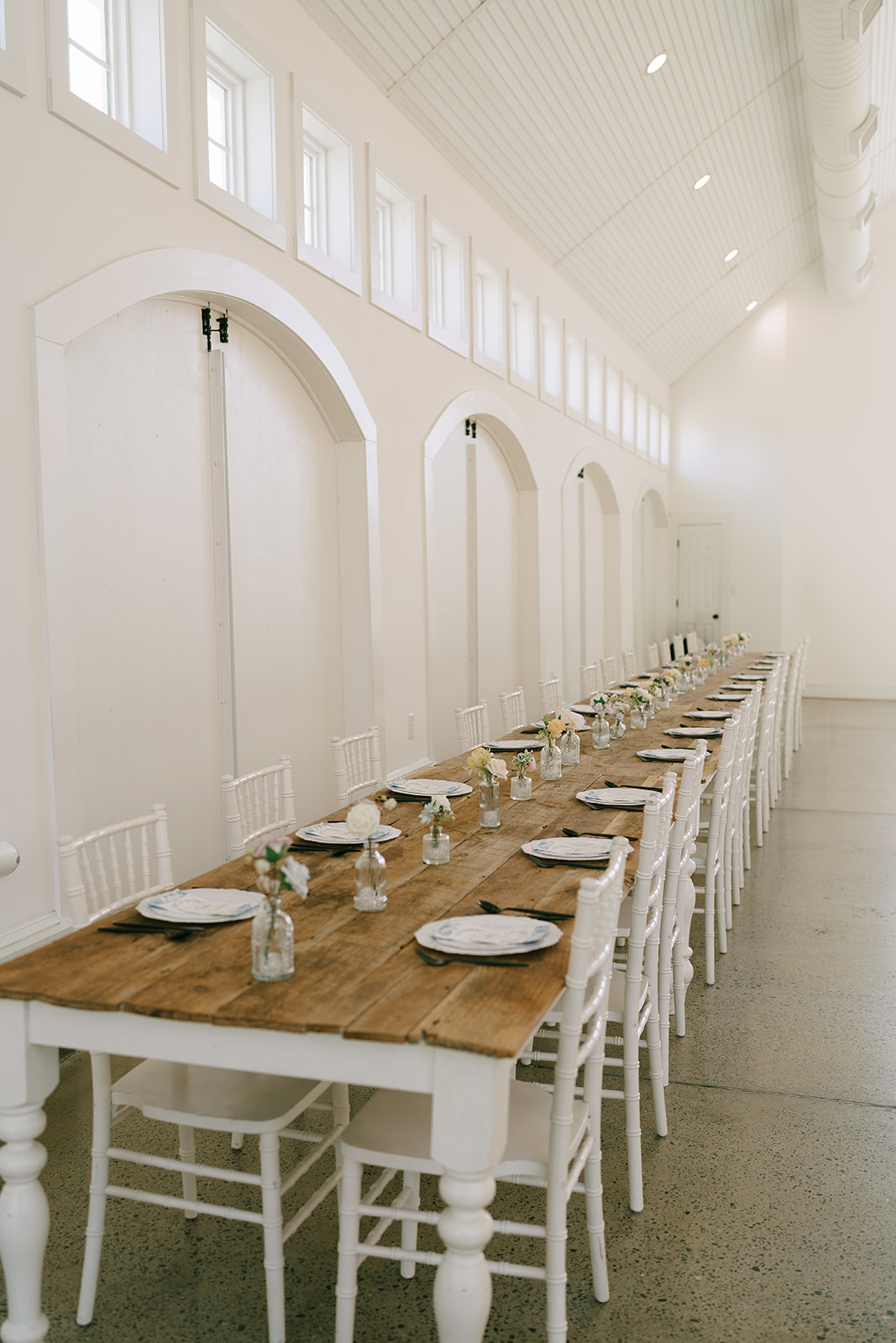 A long wooden table with white chairs is set in a bright, airy room with large windows, high ceilings, chandeliers, and minimal decor.