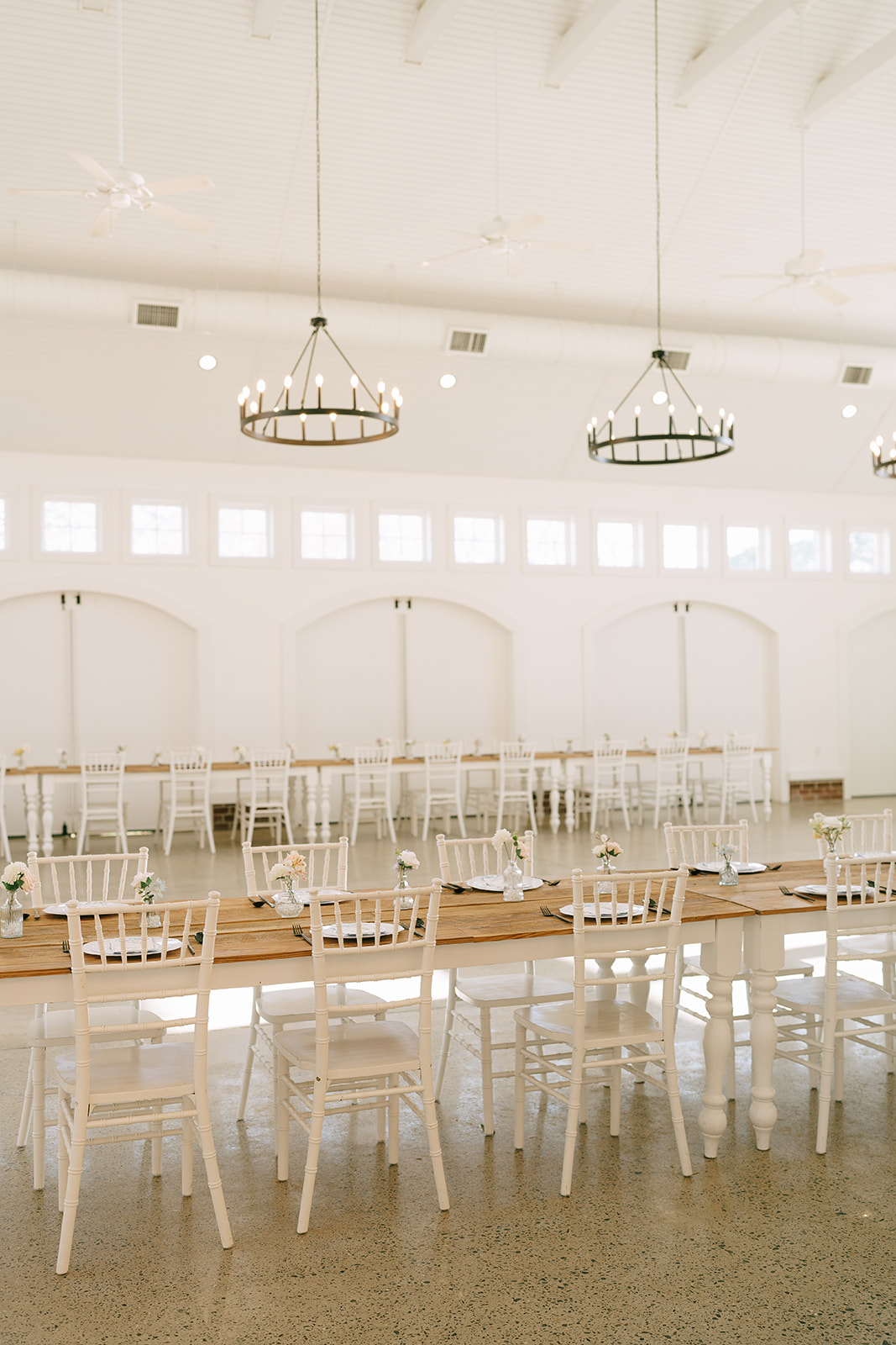 A long wooden table with white chairs is set in a bright, airy room with large windows, high ceilings, chandeliers, and minimal decor.