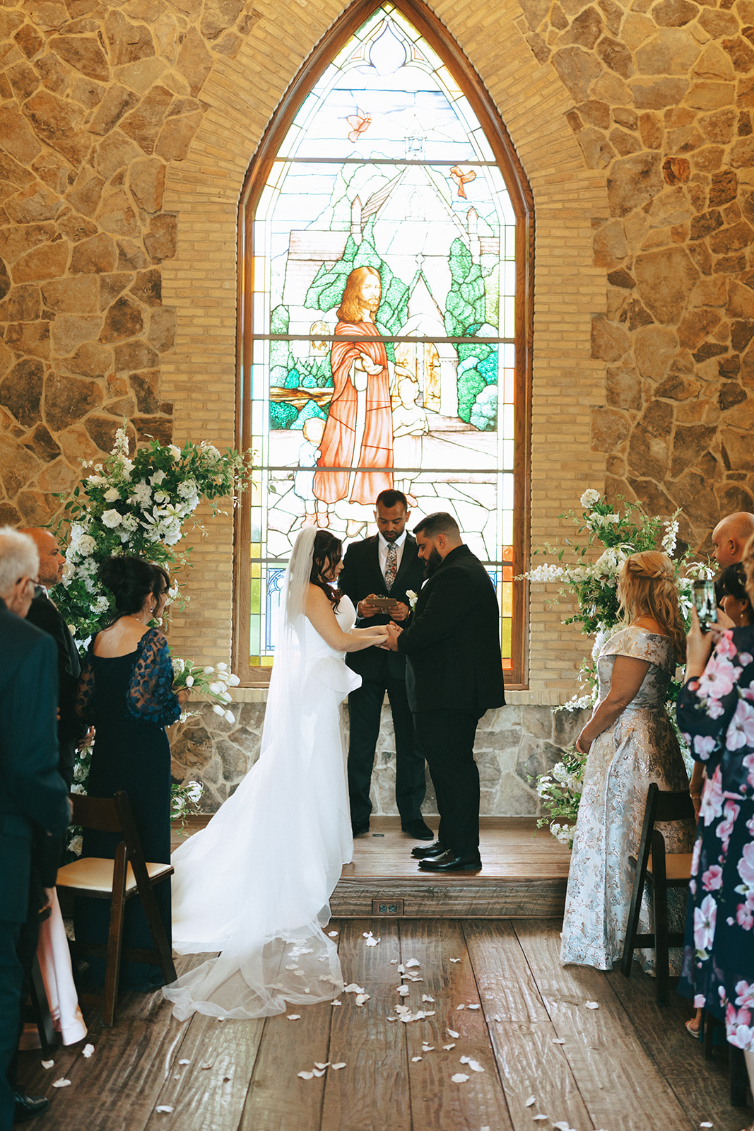 A bride and groom stand at the altar with an officiant in a stone chapel, surrounded by seated guests at the little chapel at haven ridge a wedding venue in North CarolinaA bride and groom stand at the altar with an officiant in a stone chapel, surrounded by seated guests at the little chapel at haven ridge a wedding venue in North Carolina