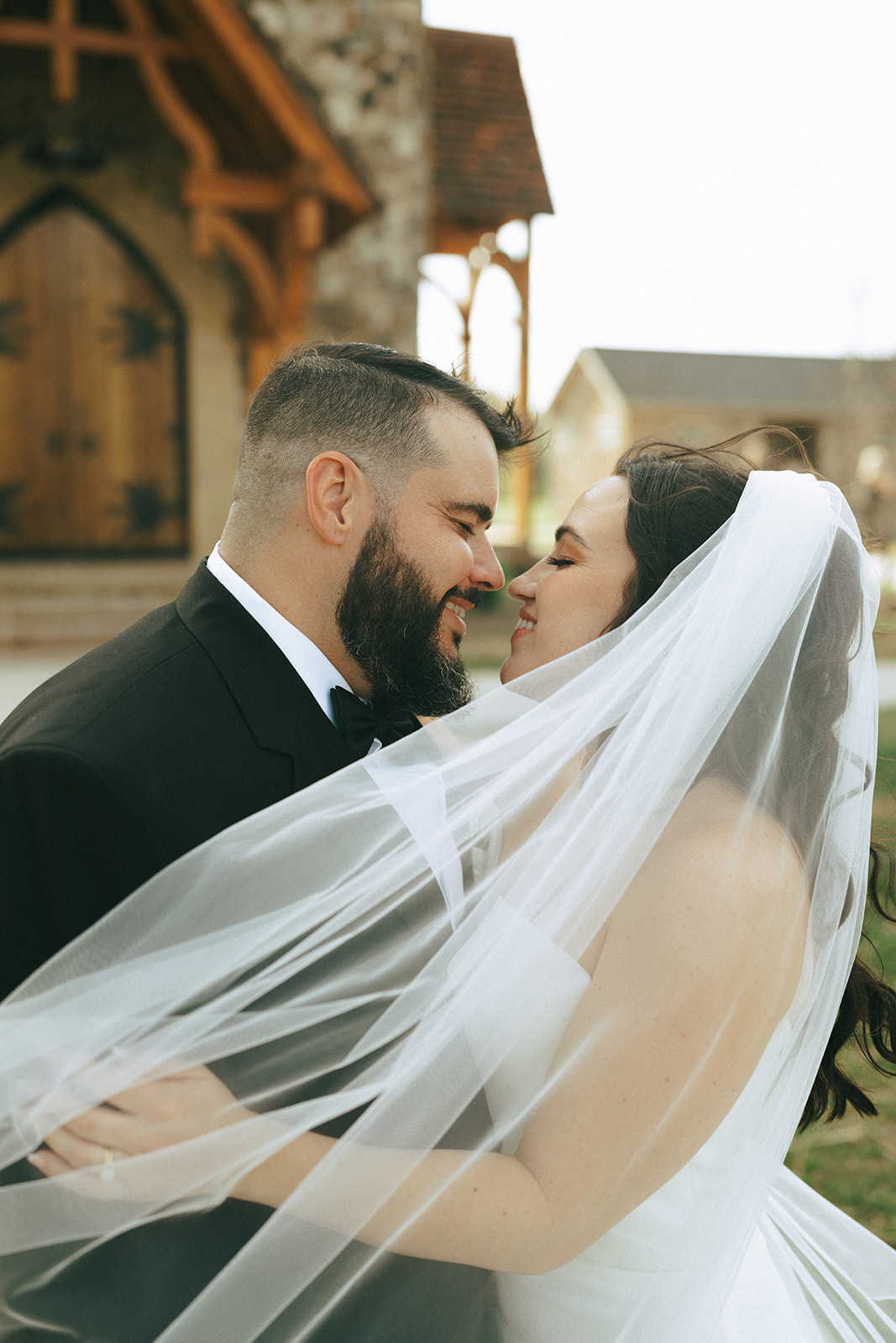 A bride and groom stand close together outdoors, smiling and touching noses, with the bride’s veil draped around them.