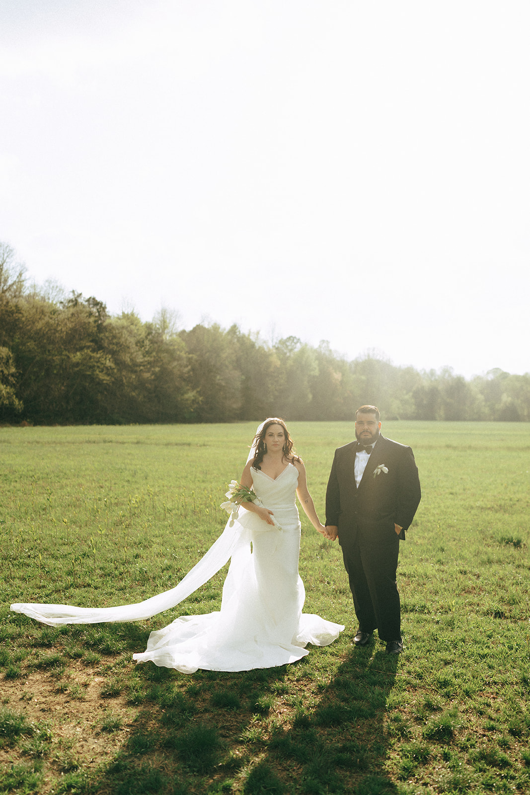 A bride in a white dress and a groom in a black suit stand together on a grassy field under bright sunlight.