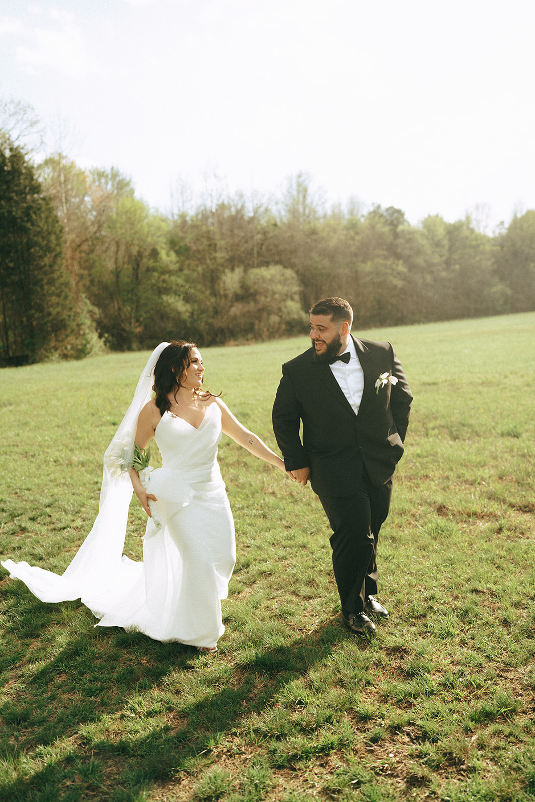 A bride in a white dress and a groom in a black suit stand together on a grassy field under bright sunlight.