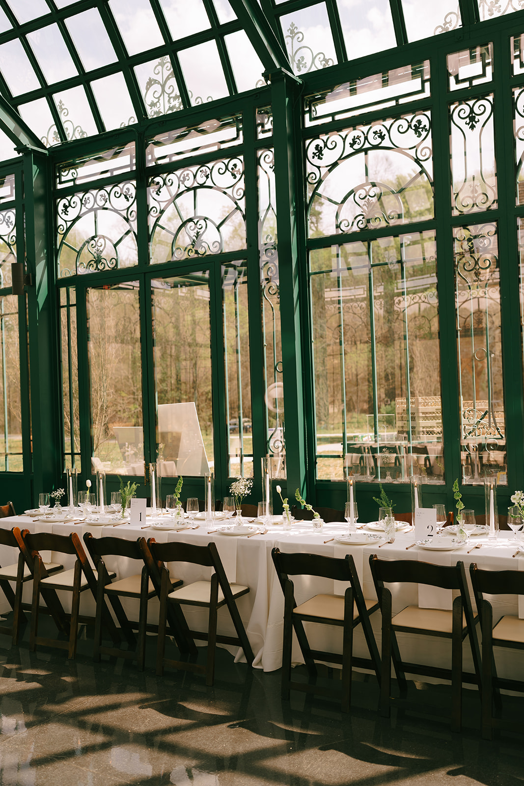 A long dining table set with white tablecloth, plates, glasses, and chairs in a sunlit glass conservatory at the little chapel at haven ridge