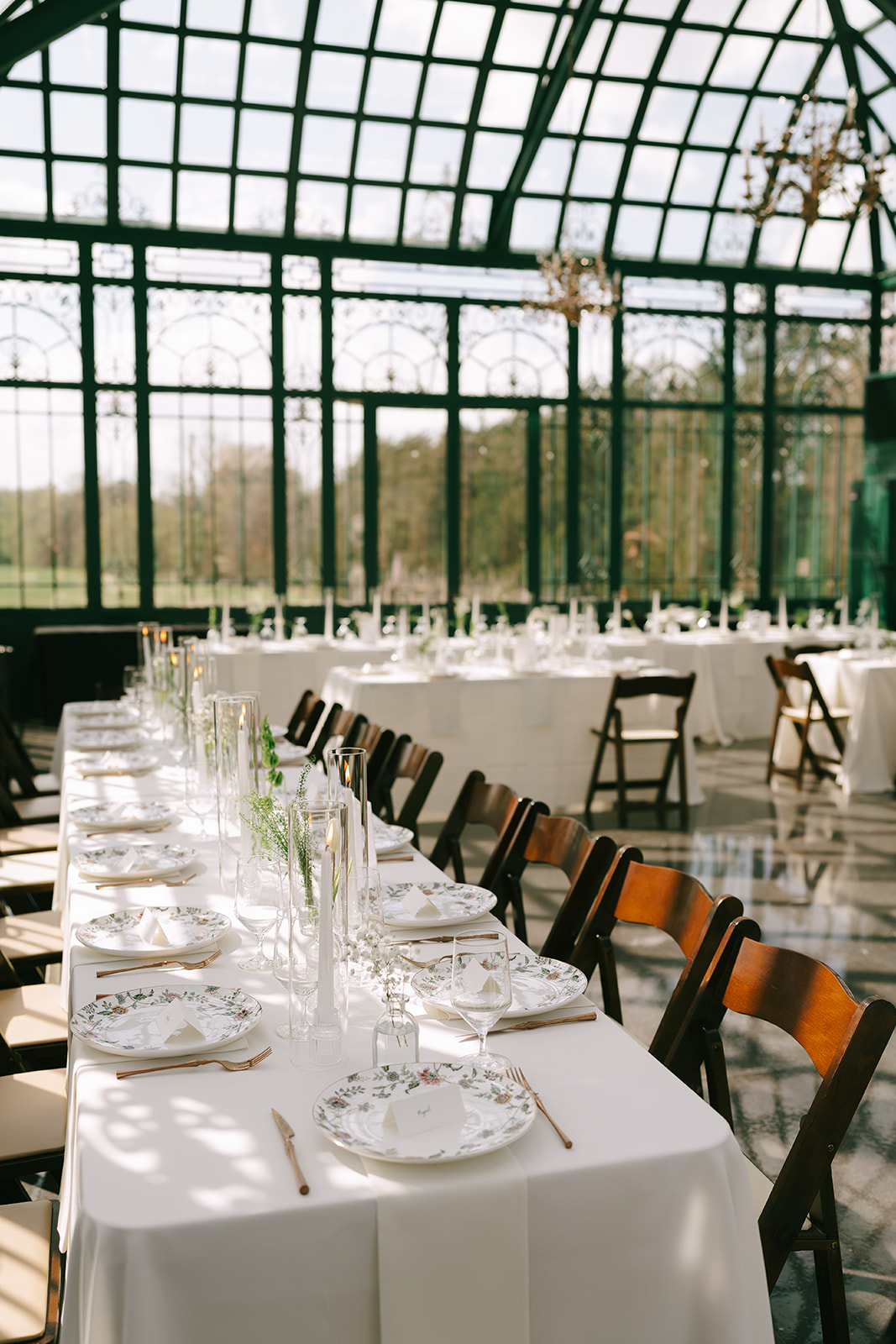 A long dining table set with white tablecloth, plates, glasses, and chairs in a sunlit glass conservatory at the little chapel at haven ridge