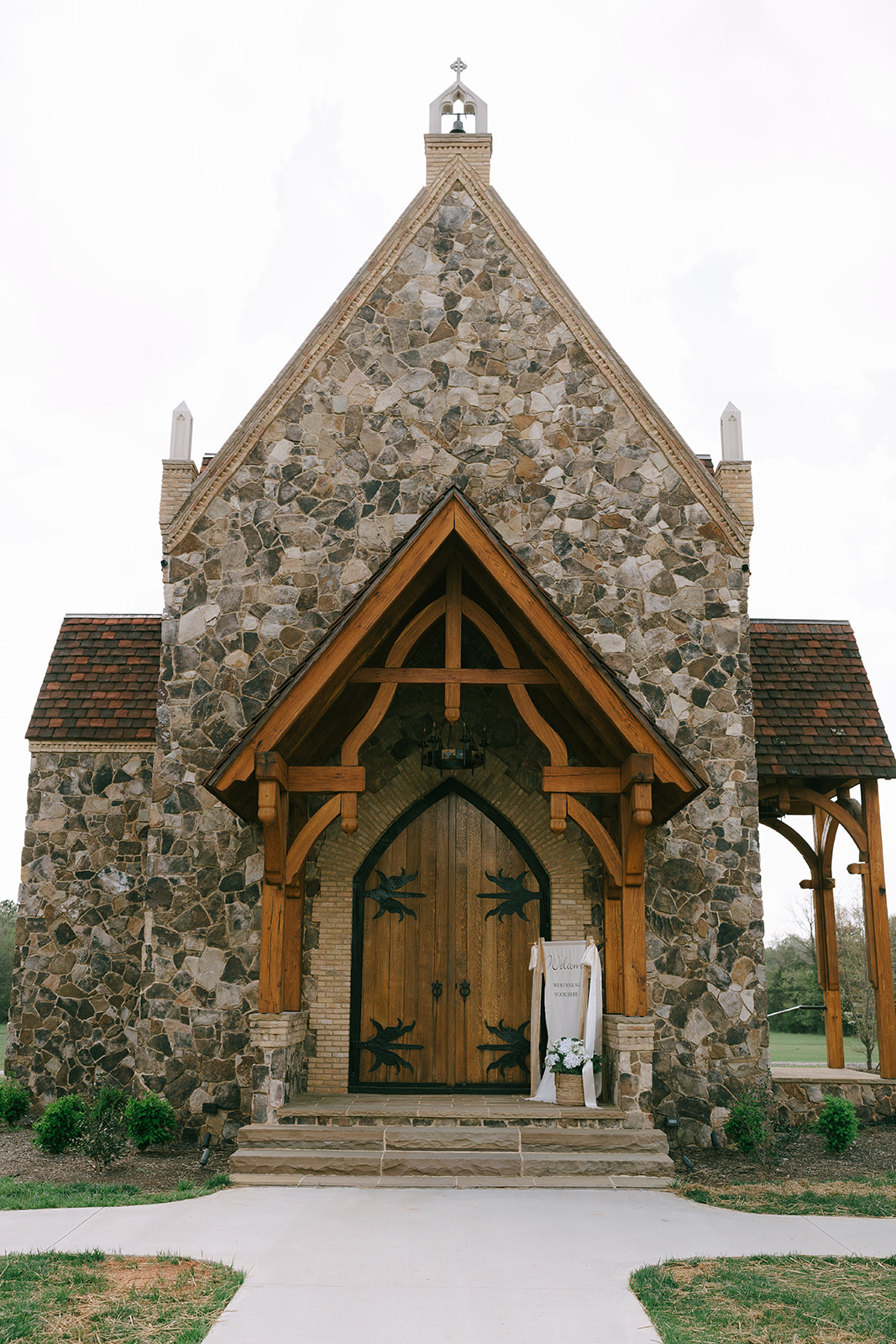 A small stone chapel with wooden double doors and a triangular roof, set on a grassy area with a concrete walkway leading to the entrance at the little chapel at haven ridge