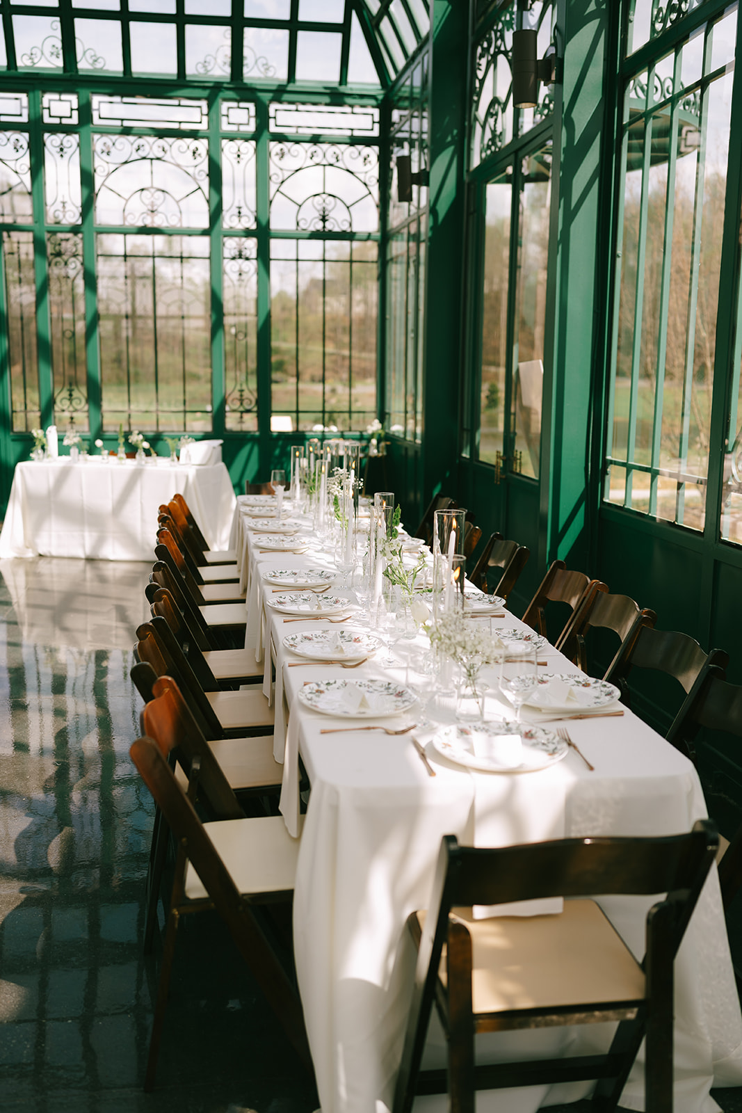 A long dining table set with white tablecloth, plates, glasses, and chairs in a sunlit glass conservatory at the little chapel at haven ridge