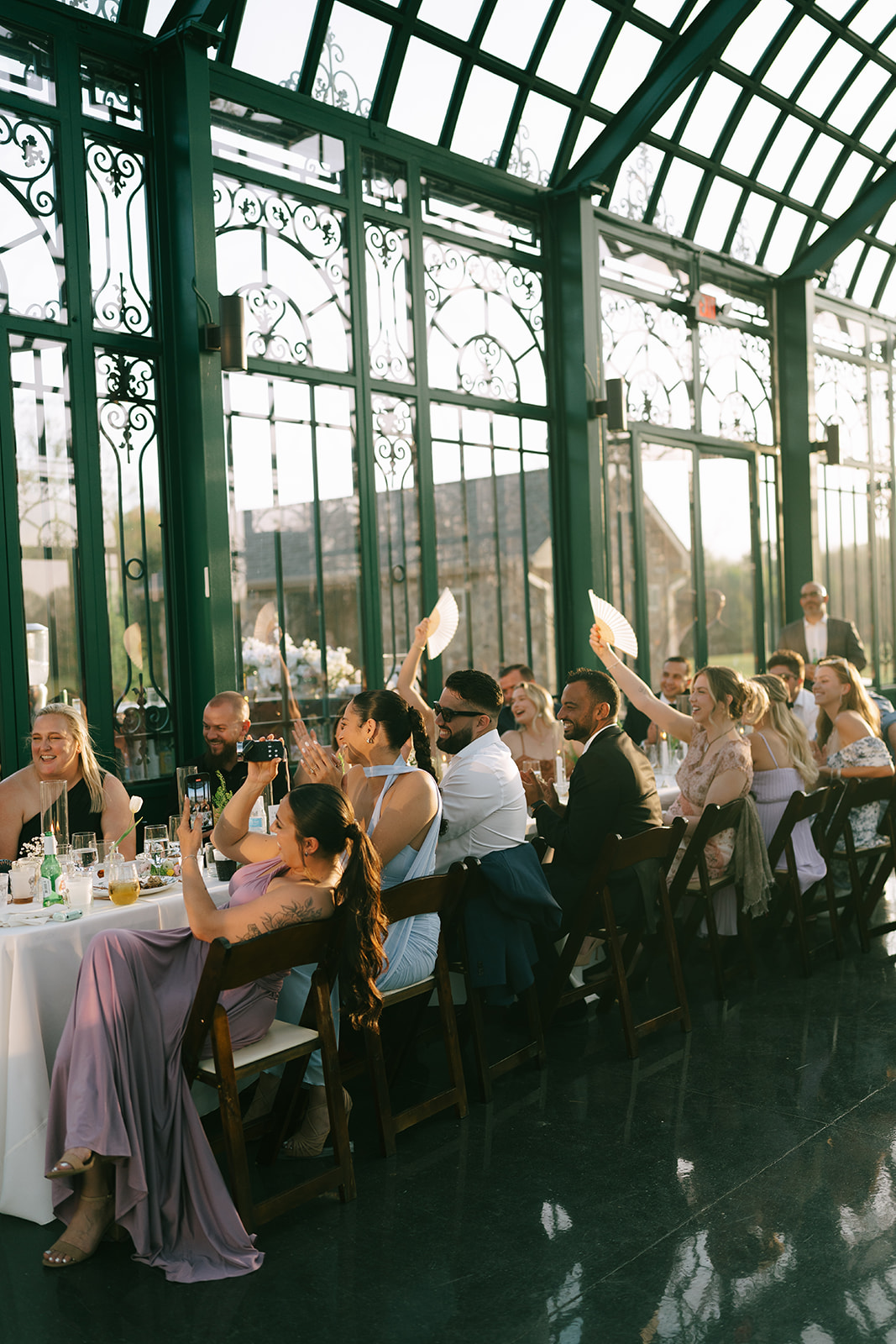 A group of people sit at a long, sunlit table set for a meal in a glass-enclosed venue, engaging in conversation.