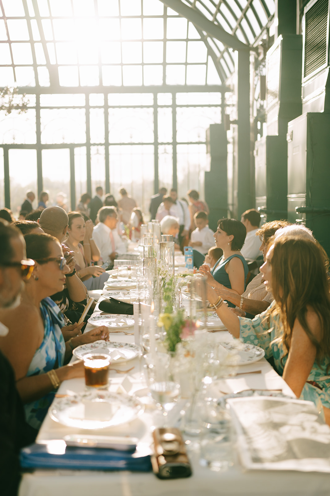 A group of people sit at a long, sunlit table set for a meal in a glass-enclosed venue, engaging in conversation.