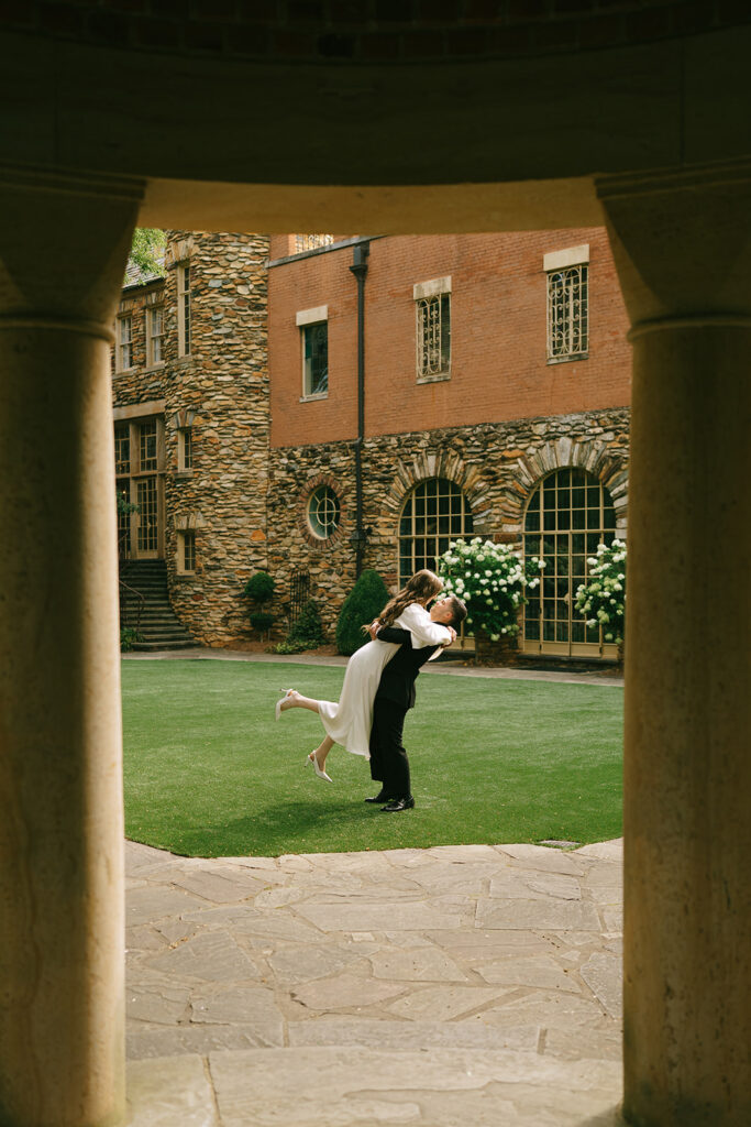 A person in a white dress is lifted by a person in a suit on a lawn, framed by stone columns with a historic brick and stone building in the background.