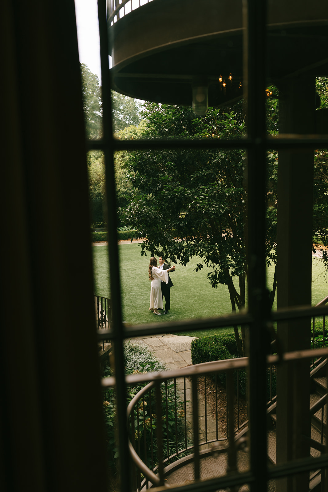Two people stand together on a lawn, seen through a window with metal bars, with trees and greenery in the background.