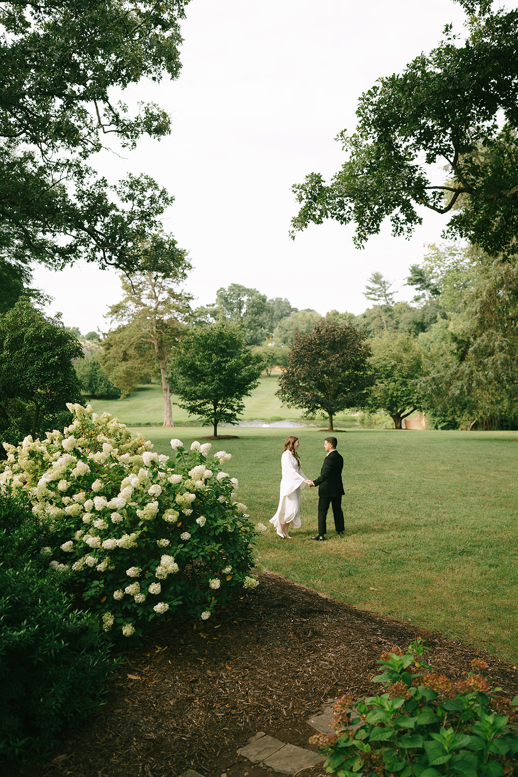 A person in a suit lifts a person in a white dress while standing in a garden with blooming white flowers at Graylyn Estate a wedding venue in North Carolina