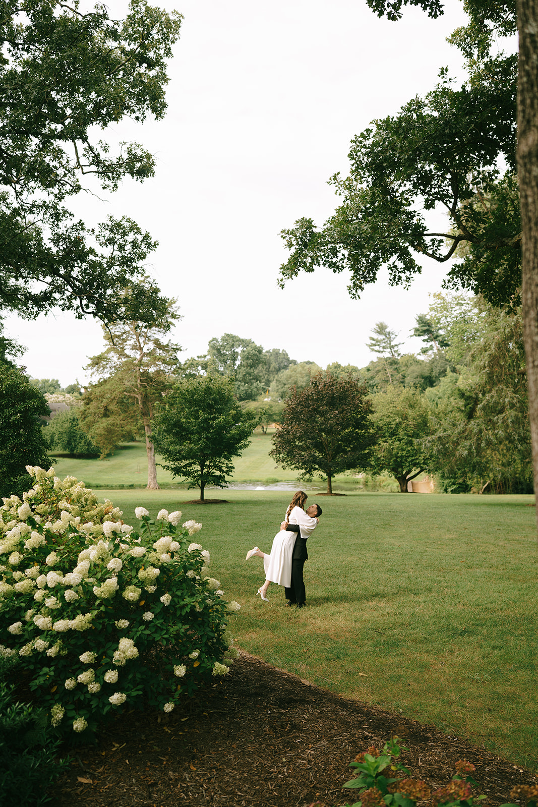 A person in a suit lifts a person in a white dress while standing in a garden with blooming white flowers at Graylyn Estate a wedding venue in North Carolina