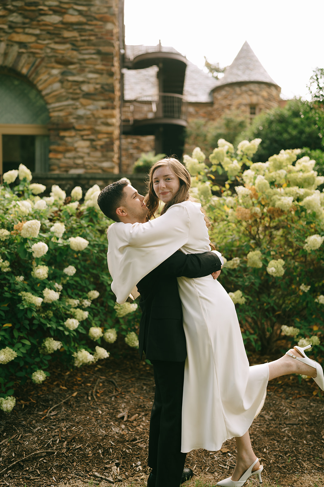 A person in a suit lifts a person in a white dress while standing in a garden with blooming white flowers and a stone building in the background at Graylyn Estate a wedding venue in North Carolina