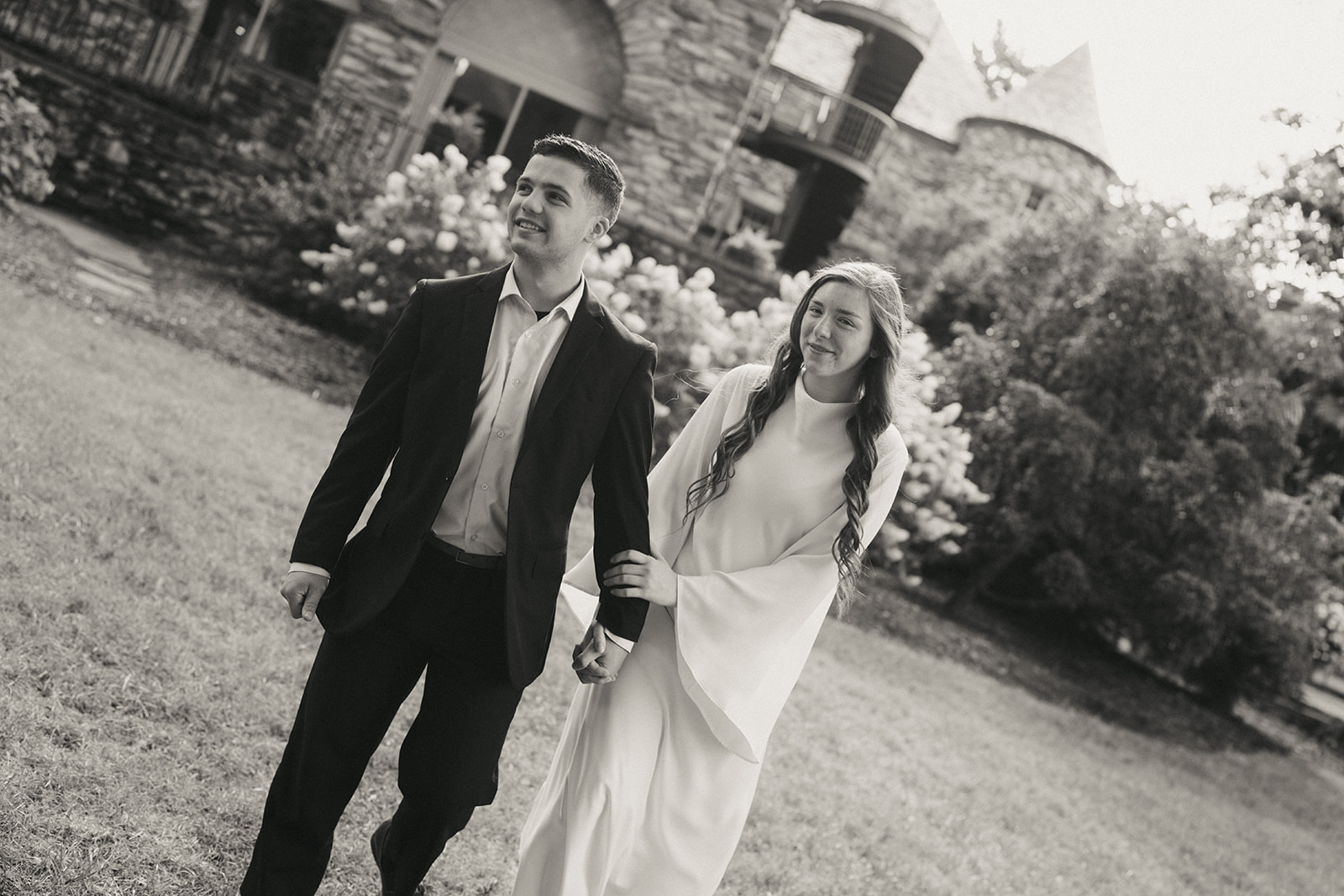 A man in a suit and a woman in a long white dress walk hand in hand outside near a stone building, both looking ahead. 
