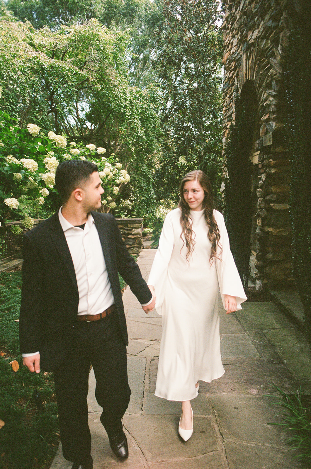 A person in a suit and a person in a white dress while standing in a garden with blooming white flowers at Graylyn Estate a wedding venue in North Carolina