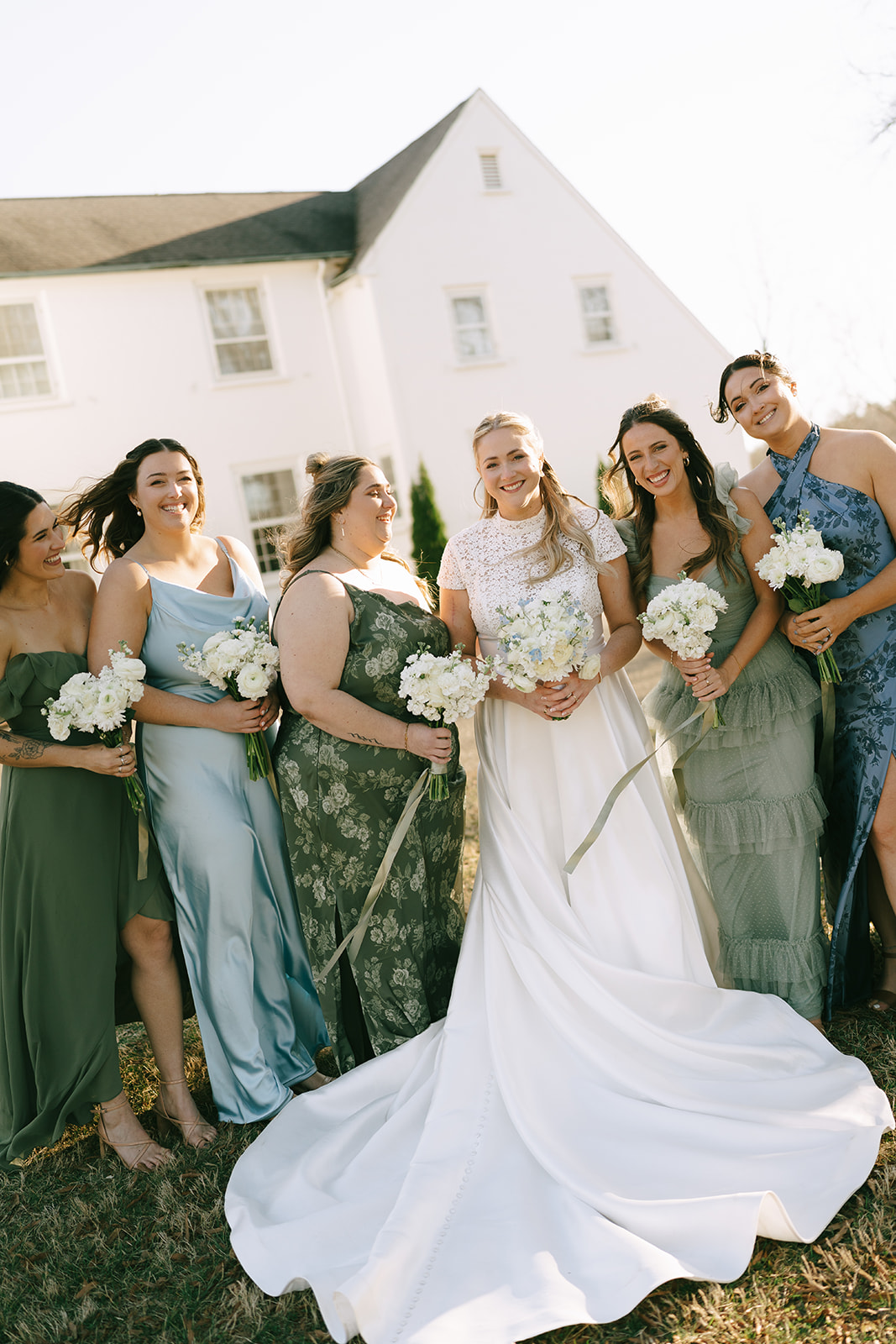 Six women in formal dresses, holding bouquets and smiling, stand closely together on a lawn in front of a large white house.