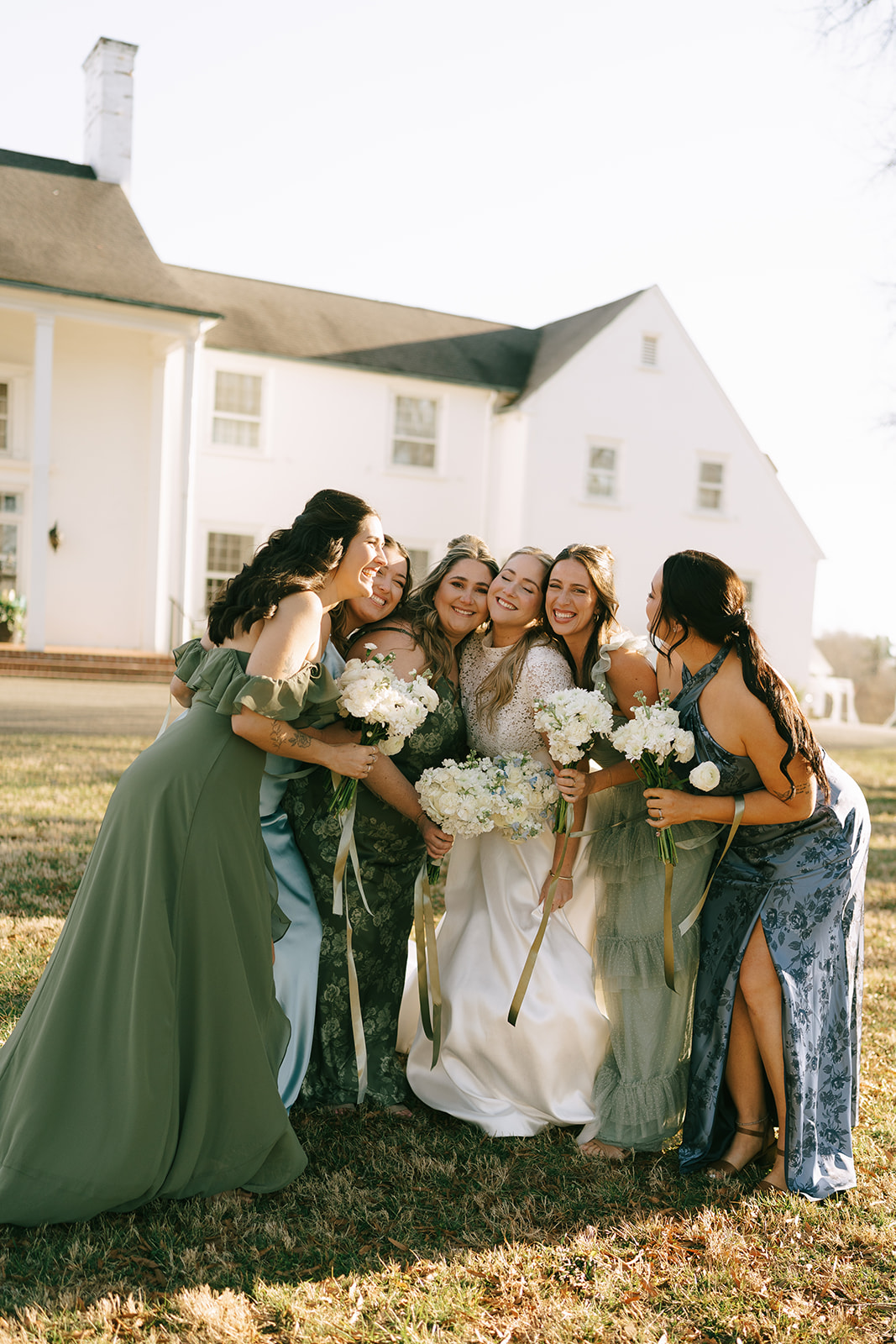 Six women in formal dresses, holding bouquets and smiling, stand closely together on a lawn in front of a large white house.