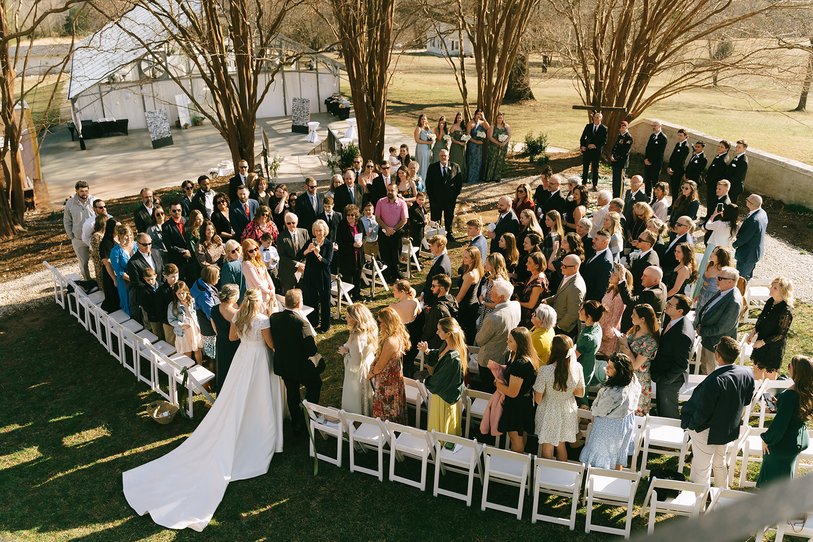 Outdoor wedding ceremony with guests seated in rows, bride and groom standing at the altar, surrounded by trees and sunlight.