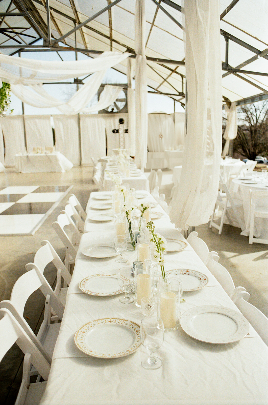 A long white table with place settings and simple floral arrangements is set for an event in a bright, sunlit indoor space with white chairs.