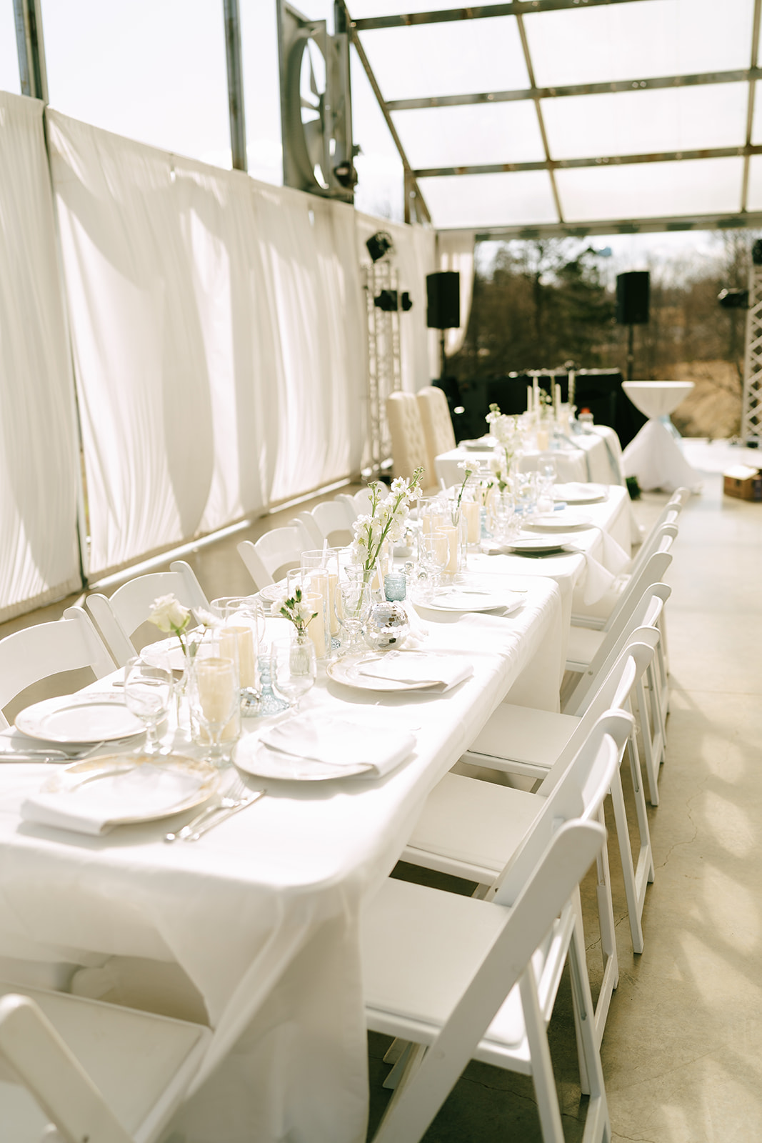 A long white table with place settings and simple floral arrangements is set for an event in a bright, sunlit indoor space with white chairs.