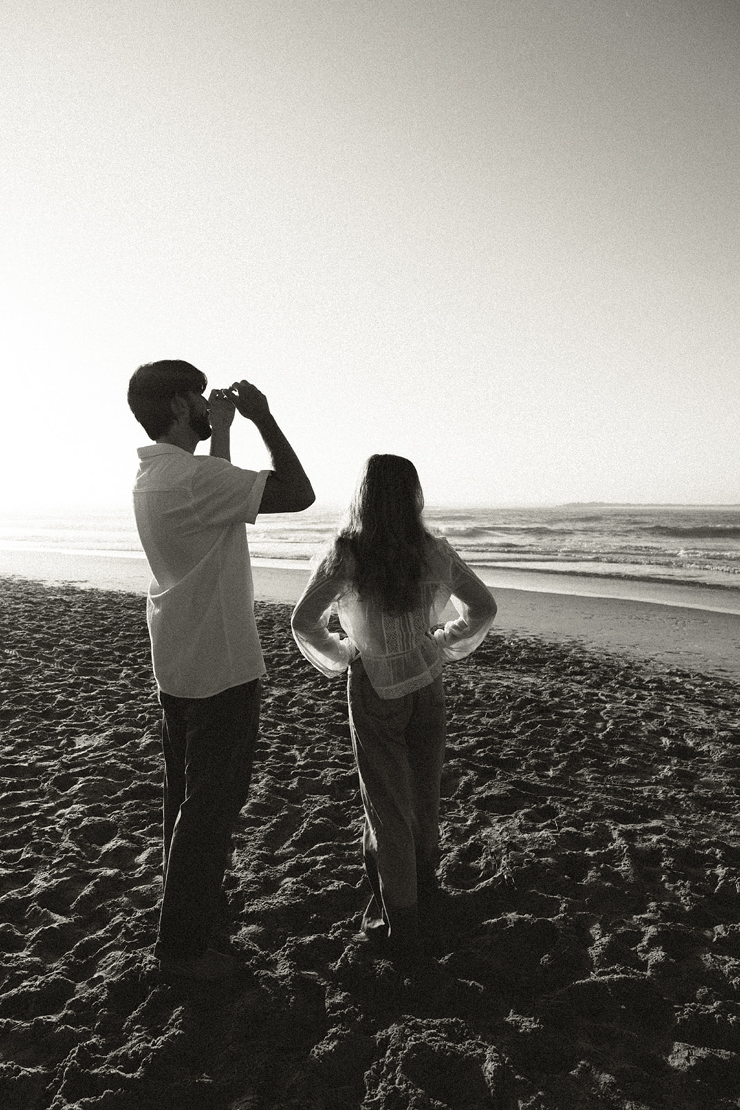 A man and woman stand on a sandy beach; the man looks through binoculars while the woman faces the ocean with her hands on her hips.
