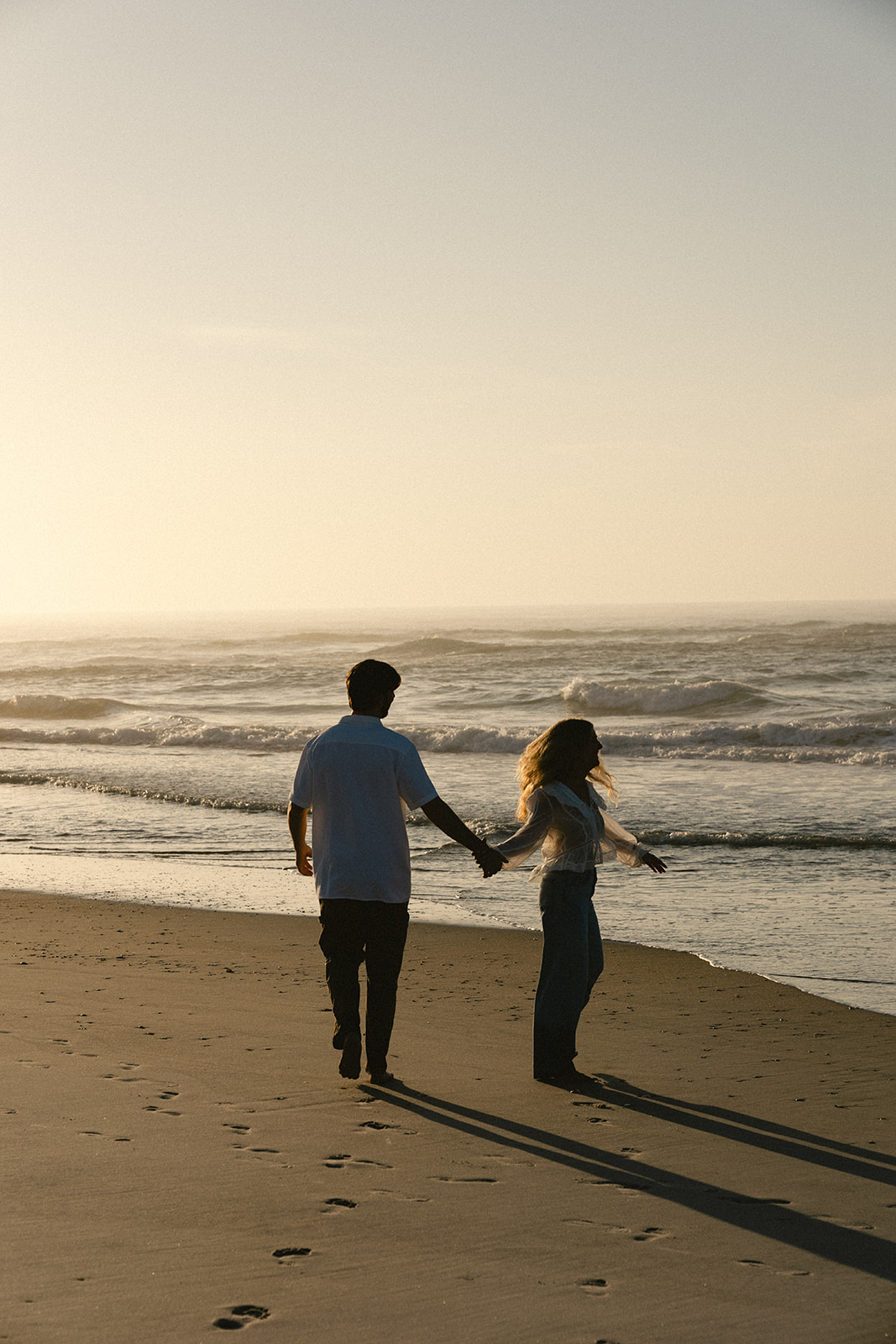 A man and woman holding hands walk along the shore at the beach during sunset, with waves in the background.