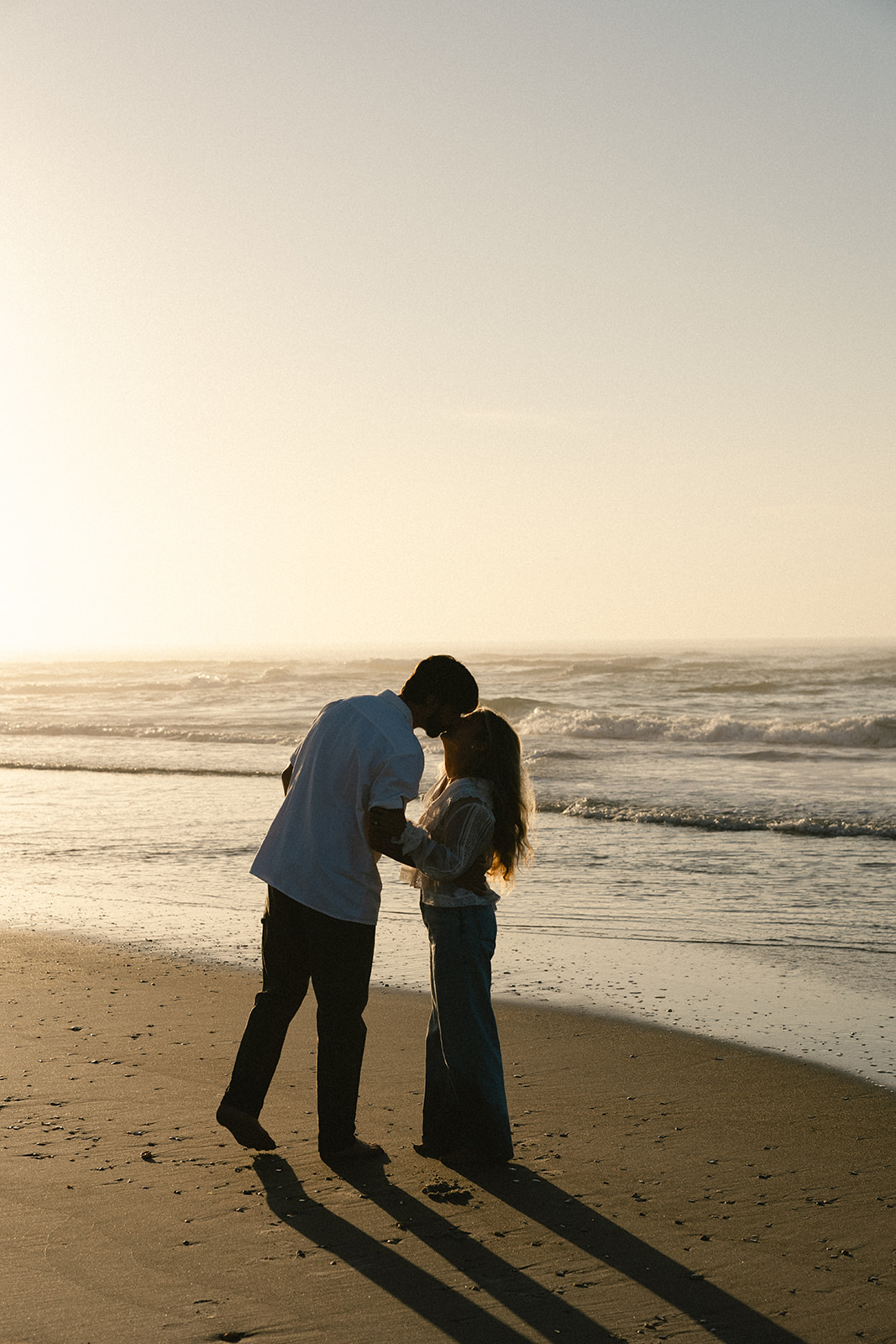 A man and woman holding hands walk along the shore at the beach during sunset, with waves in the background.