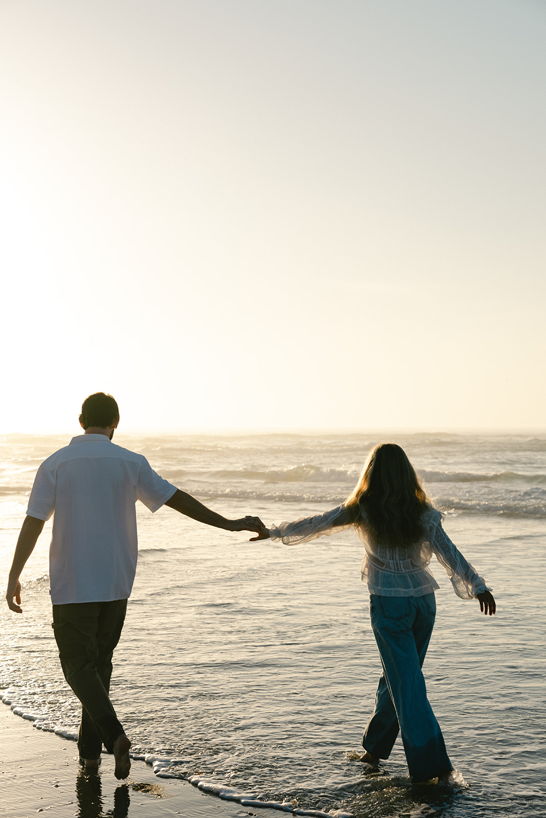A man and woman holding hands walk along the shore at the beach during sunset, with waves in the background for beach engagement photos