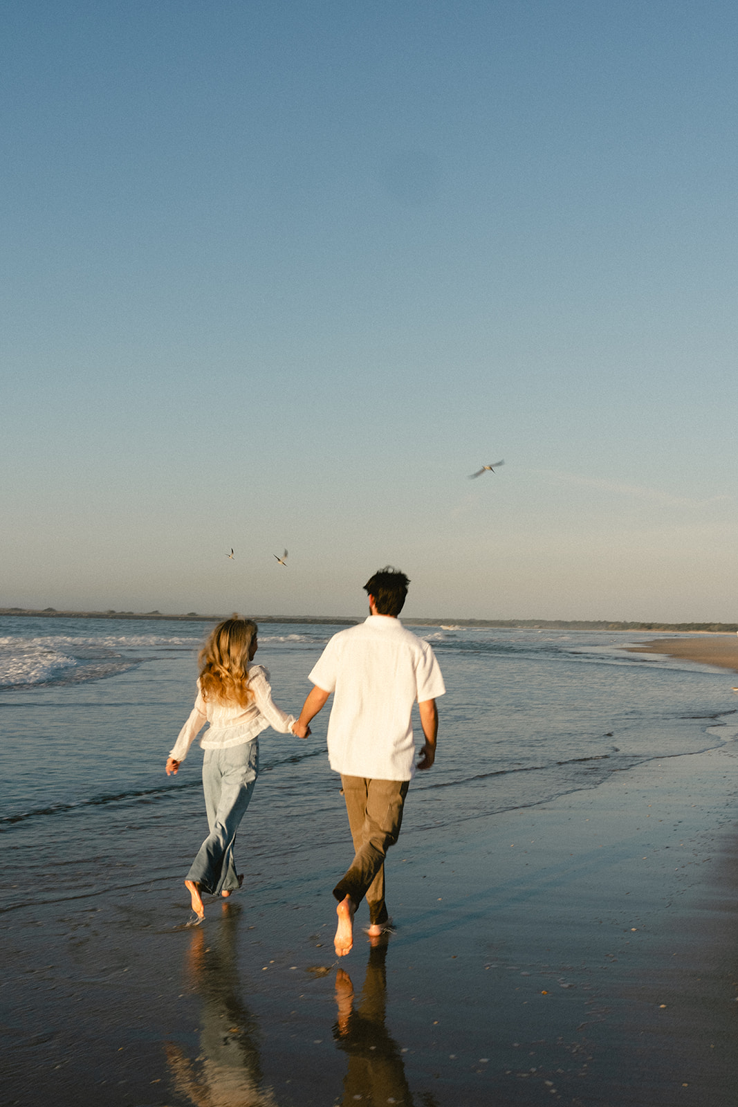 A man and woman holding hands walk along the shore at the beach during sunset, with waves in the background for beach engagement photos