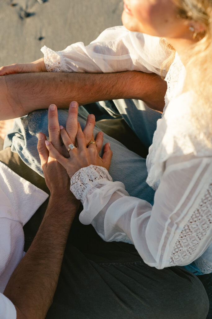 A couple sits close together on the sand, holding hands. The woman wears a white blouse and an engagement ring is visible on her finger.