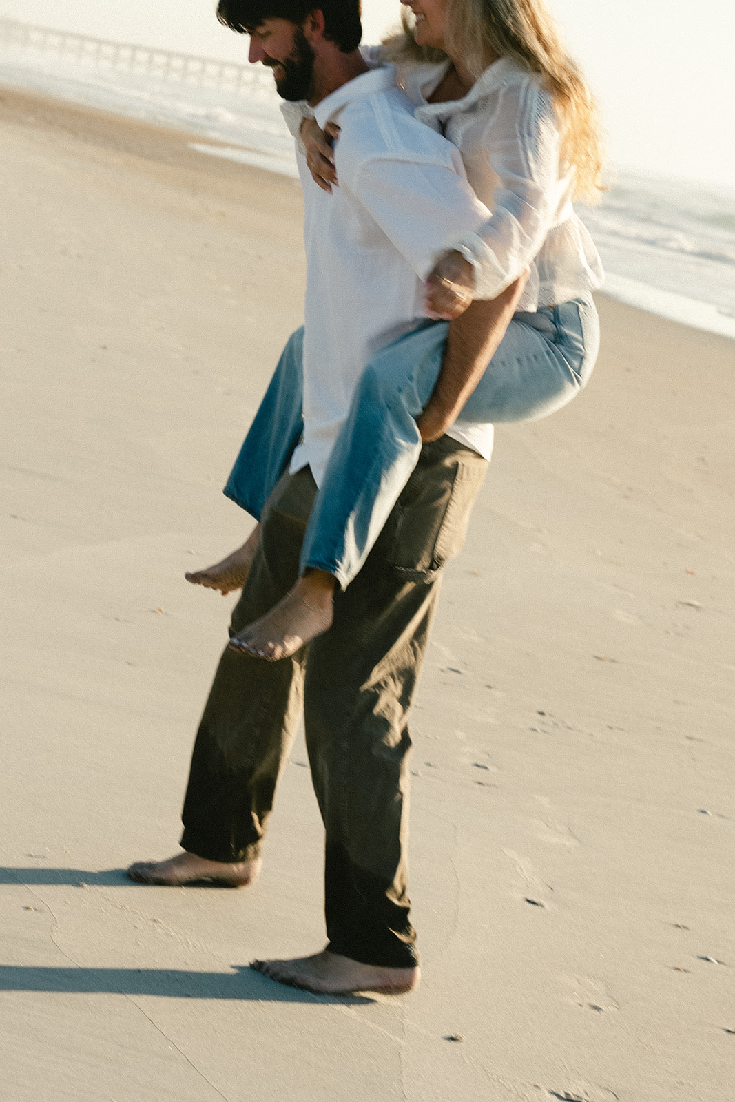 Two people stand close together at the edge of the ocean, feet in shallow water, with one person wearing light blue jeans and the other wearing dark pants.