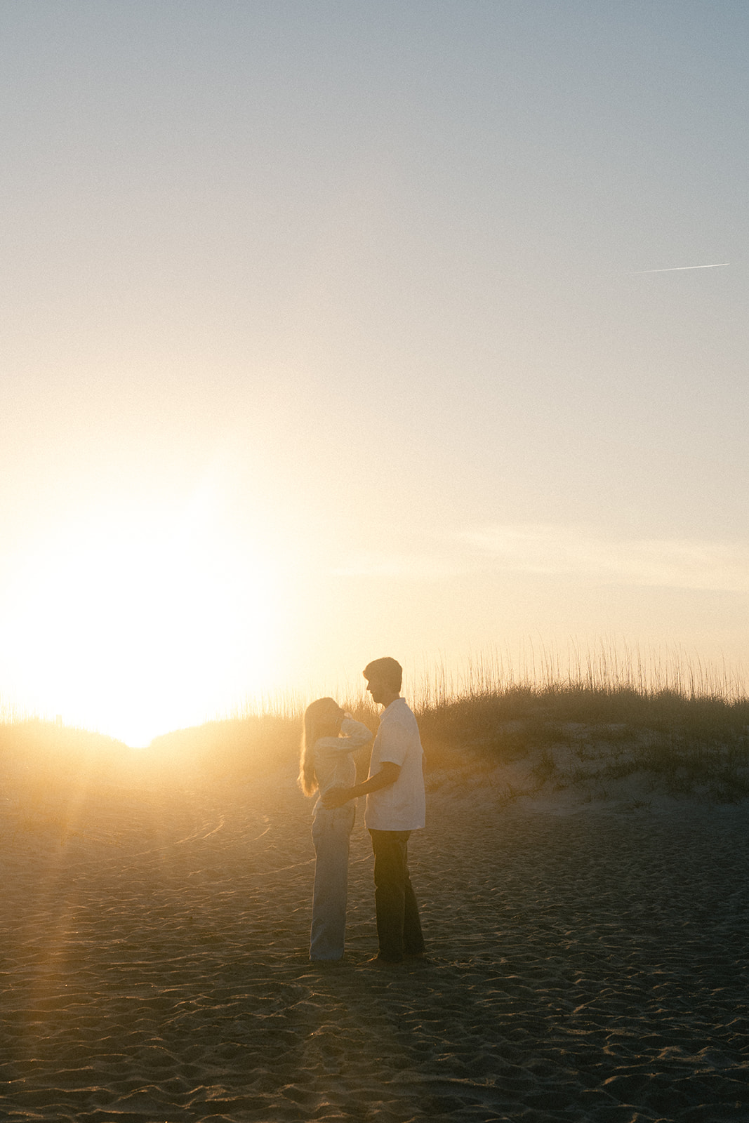 Two people stand facing each other on a sandy beach at sunset, with dunes and grasses visible in the background for their beach engagement photos