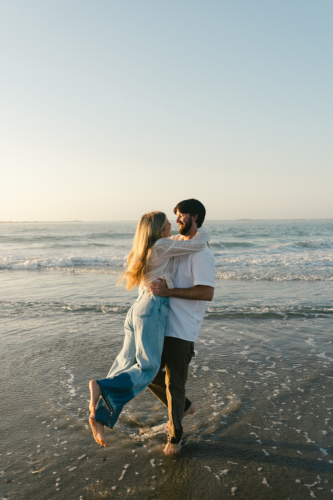 A man and woman holding hands walk along the shore at the beach during sunset, with waves in the background for beach engagement photos