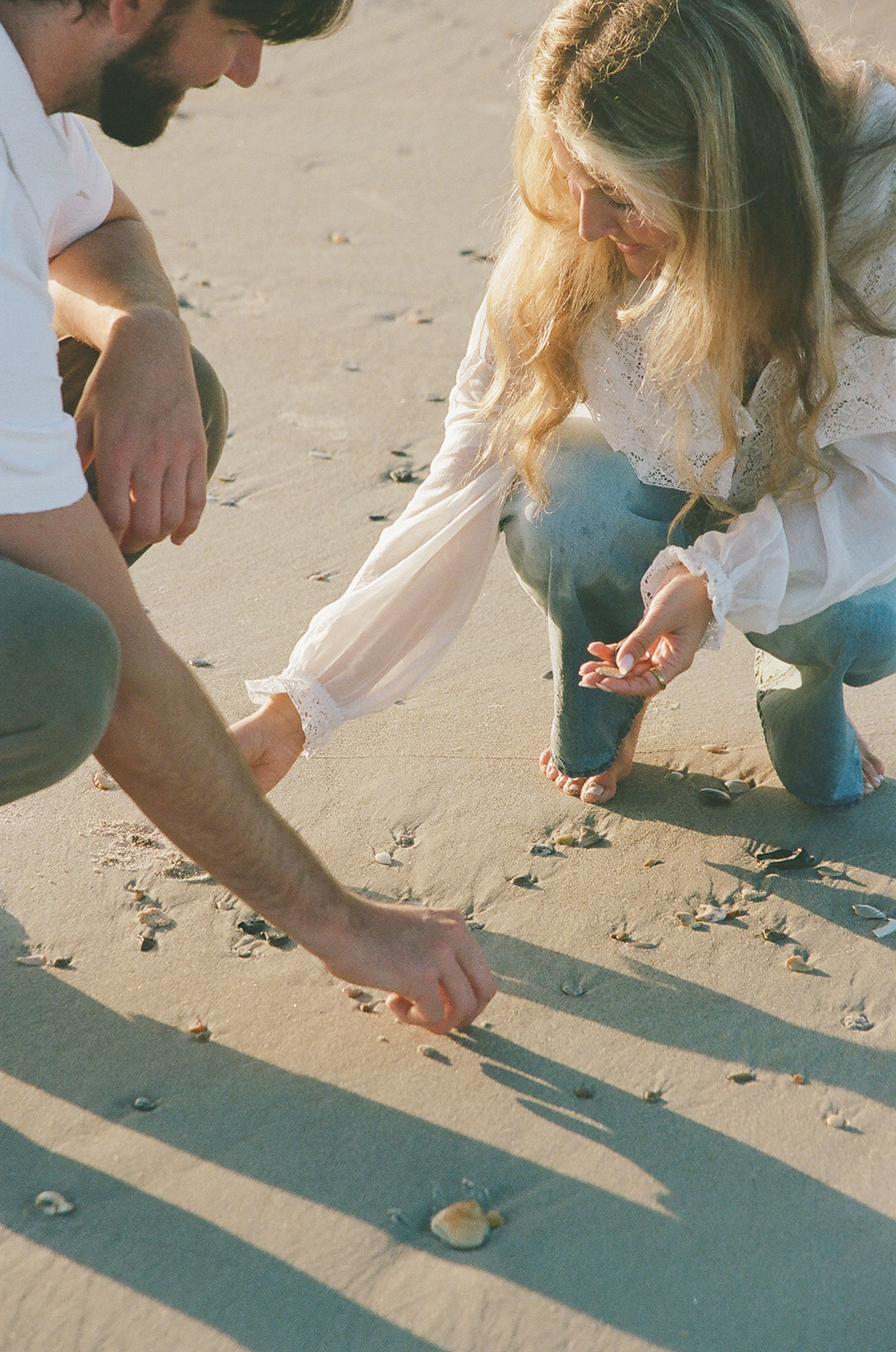 Two people crouch on a sandy beach, looking at and picking up seashells scattered on the ground.