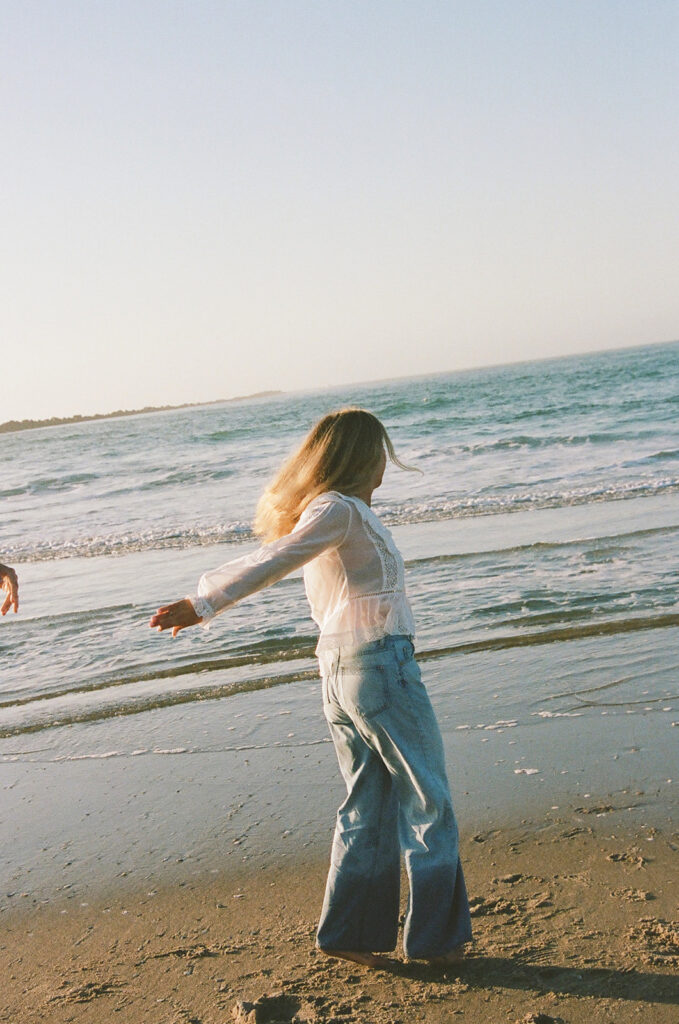 A person in a white top and blue jeans stands on a sandy beach near the shoreline with waves in the background.