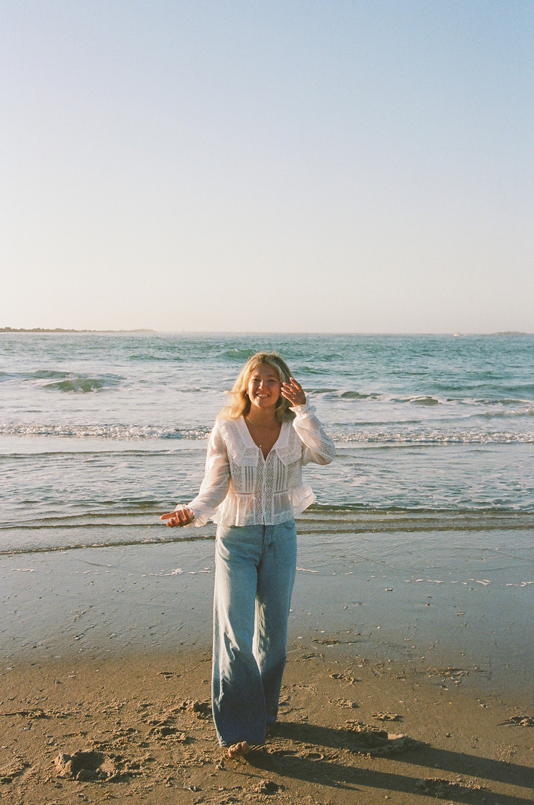 A person in a white top and blue jeans stands on a sandy beach near the shoreline with waves in the background.