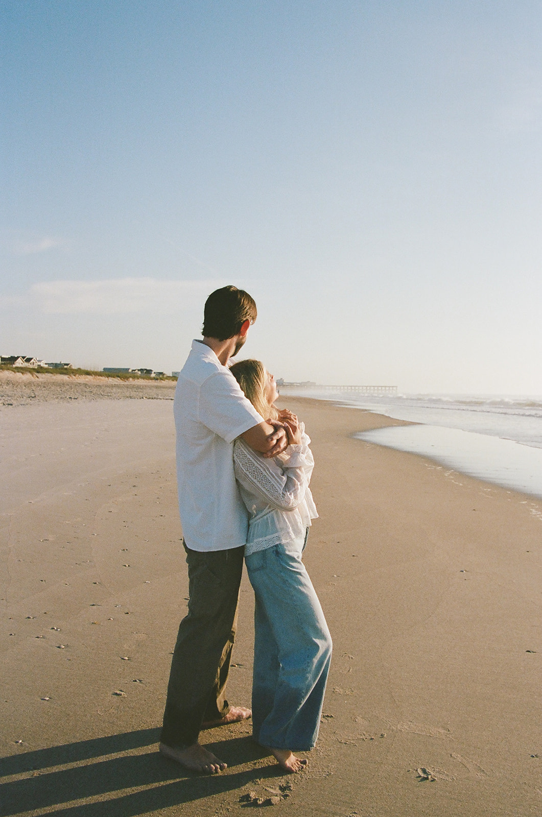 A couple stands on a sandy beach, embracing and looking out at the ocean under a clear blue sky.